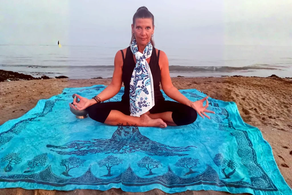 Persona seduta in meditazione a gambe incrociate su un telo con l'albero della vita, in riva al mare, con una campana accanto