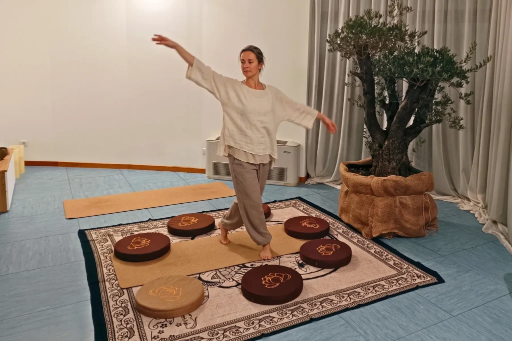 Persona in piedi su un tappeto, in equilibrio con le braccia aperte, in una sala con cuscini da meditazione e un ulivo in vaso (immagine generata con AI)
